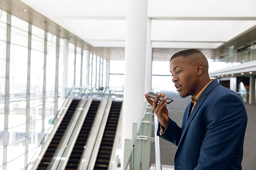 Side view close up of young African American businessman holding smartphone and speaking standing by escalators in the glass walled lobby of a modern business building. Modern corporate start up new business concept with entrepreneur working hard