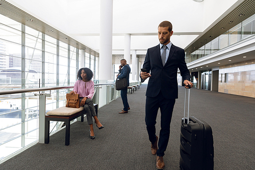 Full length front view of a young Caucasian businessman looking at smartphone while he walks wheeling a suitcase in a modern corridor. In the background an African American businesswoman is sitting on a bench and an African American businessman is talking on his phone. Modern corporate start up new business concept with entrepreneur working hard