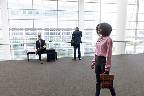 Side view of a young African American businesswoman walking holding handbag in a modern corridor while a young African American businessman stands talking on the phone and a young Caucasian businessman sits on a bench with a suitcase in the background. Modern corporate start up new business concept with entrepreneur working hard