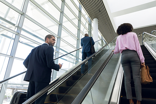 Back view of a young Caucasian businessman with suitcase holding smartphone while he stands going up on an escalator in a modern building, he looks to a young African American businesswoman going up on the next escalotor. In the background another businessman is also going up ahead of them. Modern corporate start up new business concept with entrepreneur working hard