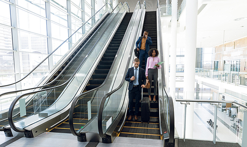 Full length front view of a young Caucasian businessman holding smartphone, a young African American businesswoman holding a file and a young African American businessman talking on a smartphone travelling down an escalator in a modern building. Modern corporate start up new business concept with entrepreneur working hard