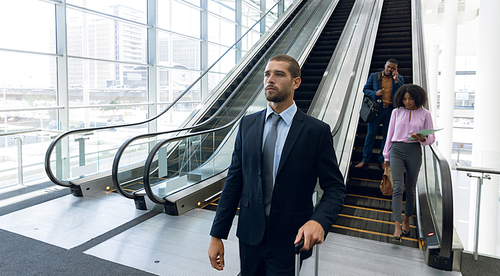 Front view of a young Caucasian businessman with a suitcase walking from an escalator in a modern building. A young African American businesswoman holding a file and a young African American businessman talking on a smartphone are travelling down the escalator behind him. Modern corporate start up new business concept with entrepreneur working hard