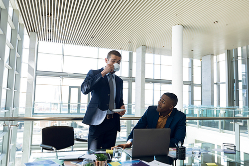 Front view of a young Caucasian businessman standing drinking coffee while a young African American businessman sits at a desk talking to him in a modern office. Modern corporate start up new business concept with entrepreneur working hard