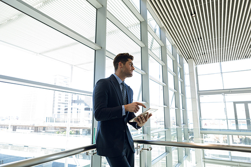 Side view of young Caucasian businessman standing using tablet computer and looking away leaning in the glass walled lobby of a modern business building. Modern corporate start up new business concept with entrepreneur working hard