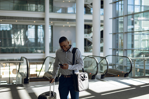 Front view of a young African American businessman with suitcase and shoulder bag using a smartphone near the escalators in a modern building. Modern corporate start up new business concept with entrepreneur working hard