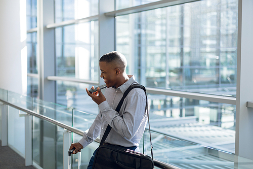 Side view of smiling young African American businessman talking on his smartphone standing in a modern building with a suitcase and a shoulder bag. Modern corporate start up new business concept with entrepreneur working hard