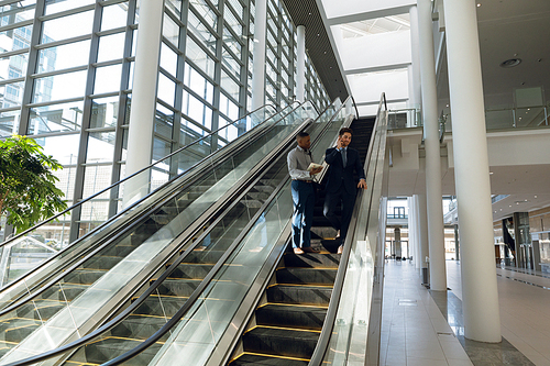 Full length view of a young African American holding documents and a young Caucasian businessman talking on smartphone both standing on an escalator in a modern building. Modern corporate start up new business concept with entrepreneur working hard