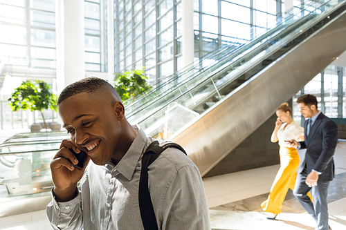 Close up side view of young African American businessman talking on smartphone by the escalators in sunlit modern business atrium. A young Caucasian businessman and businesswoman are walking together using smrtphones in the backgrouni. Modern corporate start up new business concept with entrepreneur working hard