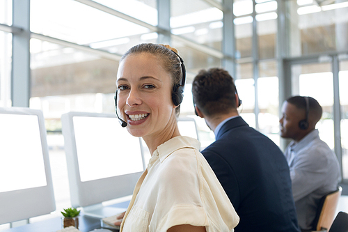 Portrait of a young Caucasian woman working in a call centre sitting at a computer wearing a headset turning to smile to camera. In the background a young Caucasian man and a young African American man are sitting at computers wearing telephone headsets working beside her in an open plan office. Modern corporate start up new business concept with entrepreneur working hard