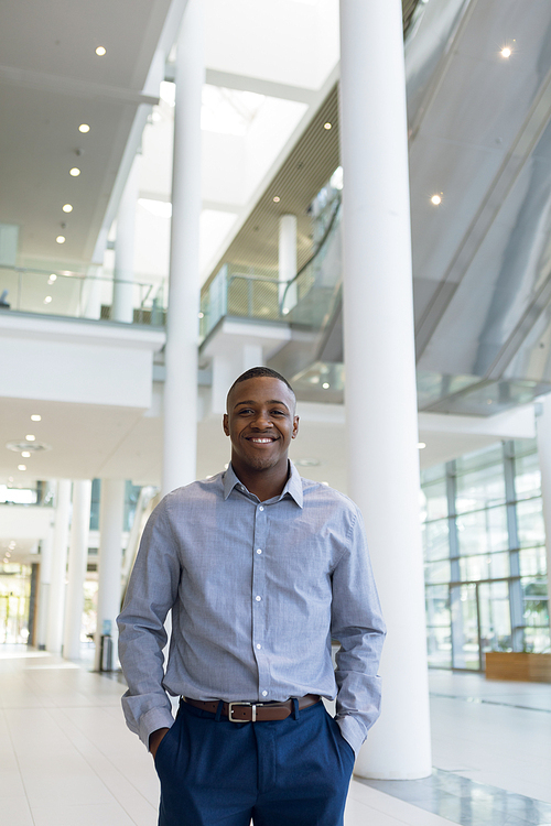 Three quarter length portrait of a smiling young African American businessman walking in the foyer of a bright modern business building with his hands in his pockets. Modern corporate start up new business concept with entrepreneur working hard