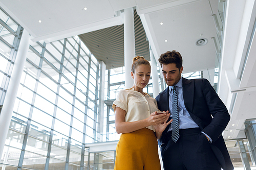 Low angle front view of a young Caucasian businesswoman and businessman looking at her smartphone in the lobby of a bright modern business building. Modern corporate start up new business concept with entrepreneur working hard