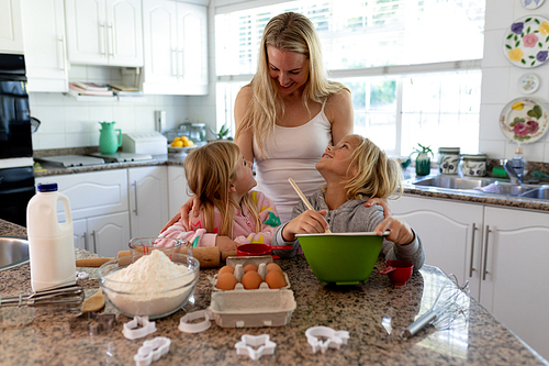 Front view of a happy young Caucasian mother with her young daughter and son in their kitchen at Christmas time making cookies, holding a mixing bowl and looking at each other