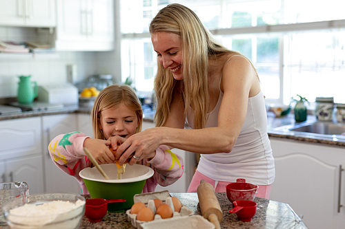 Front view of a happy young Caucasian mother with her young daughter in their kitchen at Christmas time, making cookies, cracking an egg into a mixing bowl and smiling