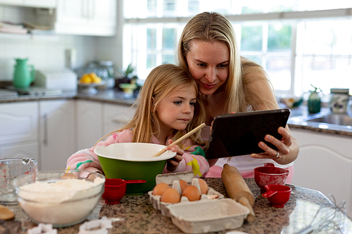 Front view of a happy young Caucasian mother with her young daughter in their kitchen at Christmas time making cookies, using a tablet and holding a mixing bowl, ingredients on the worktop in front of them