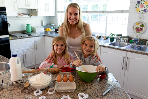 Portrait of a happy young Caucasian mother with her young daughter and son in their kitchen at Christmas time making cookies, holding a mixing bowl and smiling to camera, ingredients on the worktop in front of them