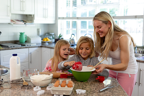 Front view of a happy young Caucasian mother with her young daughter and son in their kitchen at Christmas time making cookies, holding a mixing bowl adding ingredients and smiling