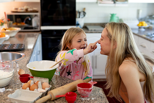Side view of a happy young Caucasian woman with her young daughter in their kitchen at Christmas time making cookies and smiling as her daughter touches her nose with flour on her finger