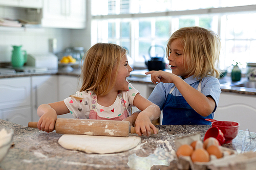 Front view of young Caucasian brother and sister in their kitchen at Christmas time making cookies, rolling dough, looking at each other and smiling