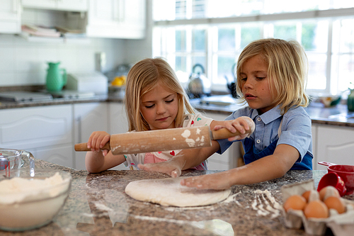 Front view of young Caucasian brother and sister in their kitchen at Christmas time making cookies, rolling dough with a rolling pin together