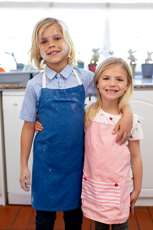 Portrait of happy young Caucasian brother and sister smiling to camera with flour on their faces and wearing aprons while making cookies in their kitchen at Christmas time, embracing and smiling