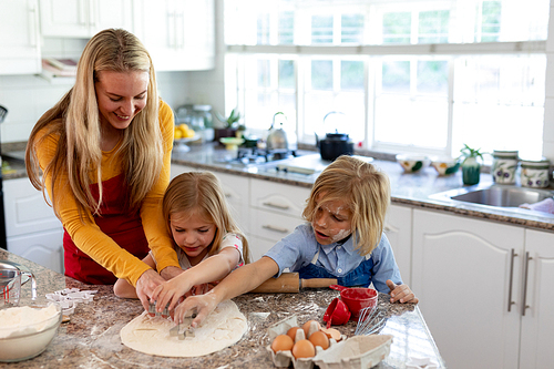 Front view of a happy young Caucasian mother with her young daughter and son in their kitchen at Christmas time making cookies, having fun using cookie cutters to cut shapes in rolled dough
