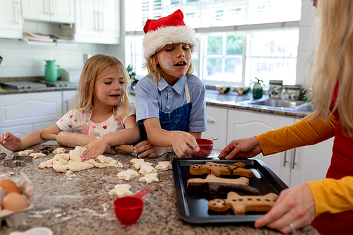 Front view of a young Caucasian mother with her young daughter and son in their kitchen at Christmas time making cookies together, mum holding a baking tray with cooked gingerbread man cookies on it, her son wearing a Santa hat
