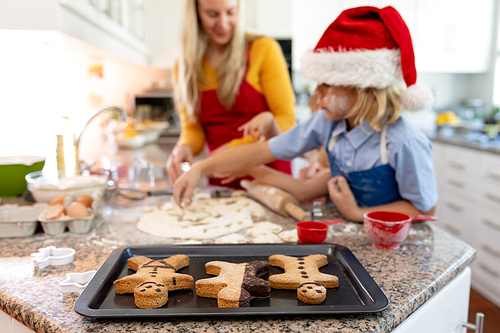 Front view of a happy young Caucasian mother with her young daughter and son in their kitchen at Christmas time making cookies, with a baking tray with cooked gingerbread man cookies on it on the worktop in the foreground, her son wearing a Santa hat