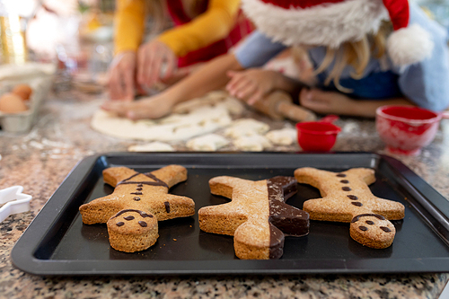 Front view close up of a young Caucasian mother with her young daughter and son in their kitchen at Christmas time making cookies, with a baking tray with cooked gingerbread man cookies on it on the worktop in the foreground