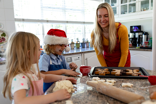 Front view of a happy young Caucasian mother with her young daughter and son in their kitchen at Christmas time making cookies, the children rolling dough, and a baking tray with cooked gingerbread man cookies on it on the worktop