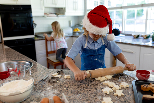 Front view of young Caucasian boy rolling dough on a worktop with his sister in the background in their kitchen at Christmas time while making cookies
