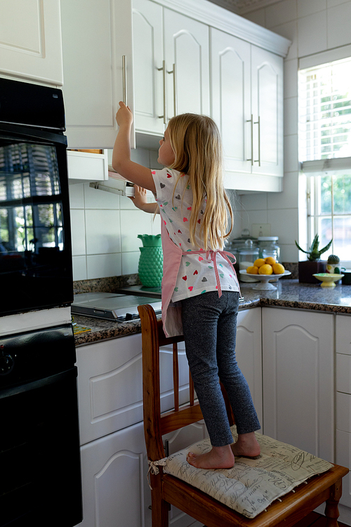 Side view of a young Caucasian girl in her kitchen standing on a chair and looking in a kitchen cabinet