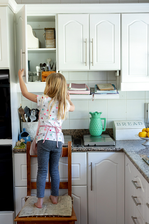 Rear view of a young Caucasian girl in her kitchen standing on a chair and looking in a kitchen cabinet