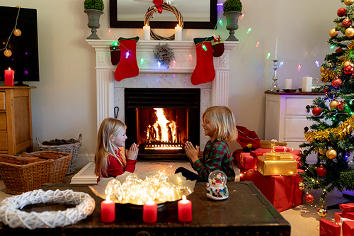 Side view of young Caucasian brother and sister sitting by the fireplace in their sitting room at Christmas time saying prayers