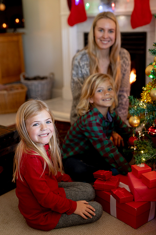 Portrait of a young Caucasian woman and her young son and daughter sitting on the floor in their sitting room at Christmas time beside a decorated Christmas tree and wrapped presents smiling to camera