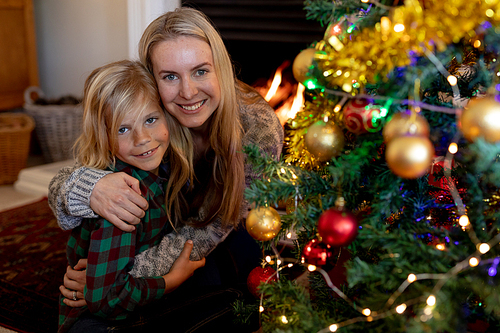 Front view of a young Caucasian woman embracing her young son sitting on the floor in their sitting room at Christmas time beside a decorated Christmas tree, smiling to camera