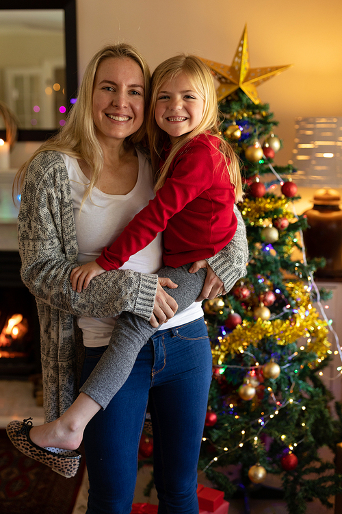 Front view of a young Caucasian woman holding her young daughter in their sitting room at Christmas time beside a decorated Christmas tree smiling to camera