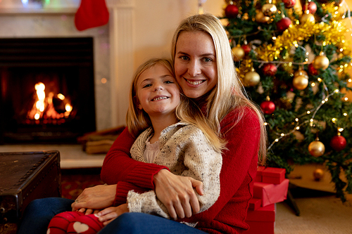 Front view of a young Caucasian woman embracing her young daughter on her knee in their sitting room at Christmas time beside a decorated Christmas tree, smiling to camera