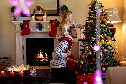 Side view of a middle aged Caucasian man with his young daughter sitting on his shoulders decorating the Christmas tree in their sitting room, with Christmas star decorations hanging in the foreground