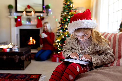 Front view close up of a young Caucasian girl wearing a Santa hat sitting on the sofa using a tablet in her sitting room at Christmas time, with her mother sitting by the fireplace in the background