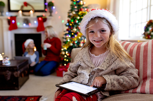 Front view close up of a young Caucasian girl wearing a Santa hat sitting on the sofa using a tablet in her sitting room at Christmas time smiling to camera, with her mother and brother sitting by the fireplace in the background