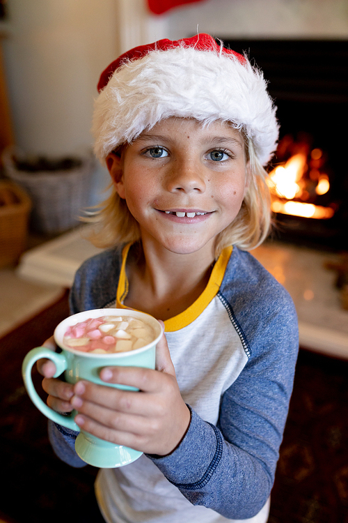 Front view of a young Caucasian boy wearing a Santa hat holding a cup of hot chocolate with marshmallows in his sitting room at Christmas time, smiling to camera