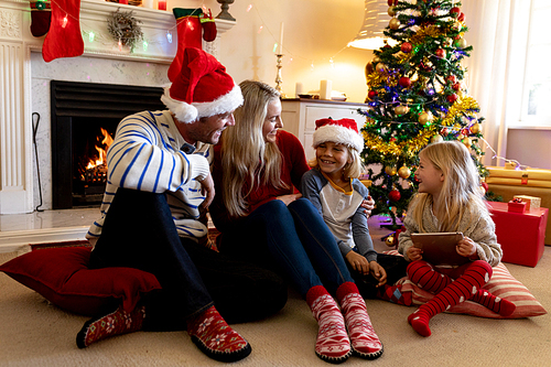 Front view of a Caucasian couple with their young son and daughter sitting on the floor in their sitting room at Christmas time, wearing Santa hats, talking and smiling to each other, the girl is holding a tablet computer