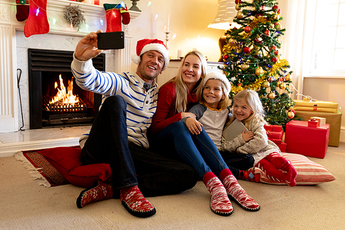 Front view of a Caucasian couple with their young son and daughter sitting on the floor in their sitting room at Christmas time, dad taking a selfie with his smartphone