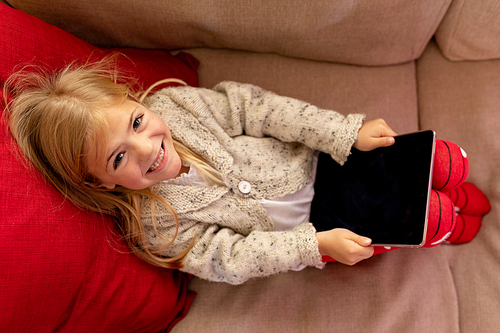 High angle view of a happy young Caucasian girl holding a tablet computer and reclining on a sofa in her sitting room at Christmas time, smiling to camera