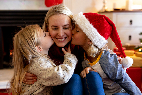 Front view close up of a young Caucasian woman sitting on the floor between her young son and daughter in their sitting room at Christmas time, the children kissing her on either cheek