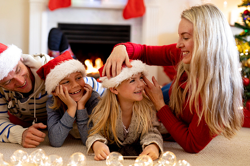 Front view of a Caucasian couple with their young son and daughter, lying on the floor wearing Santa hats in their sitting room at Christmas time, mother adjusting the hat on her daughter