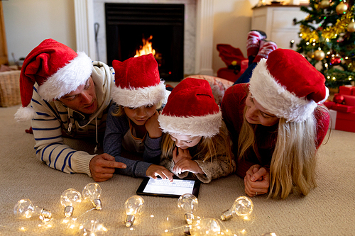 Front view of a Caucasian couple lying on the floor with their young son and daughter in their sitting room at Christmas time wearing Santa hats and using a tablet computwe together