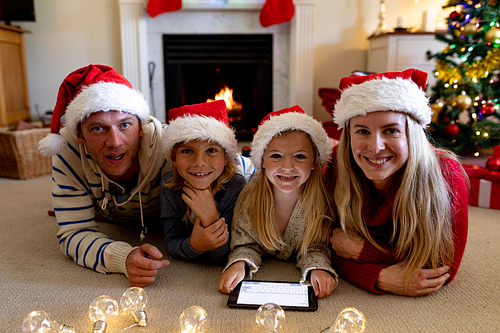 Portrait of a Caucasian couple lying on the floor with their young son and daughter smiling to camera in their sitting room at Christmas time, wearing Santa hats and using a tablet computer