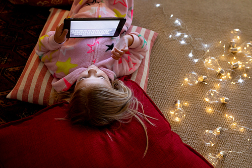 Front view of a happy young Caucasian girl lying on cushions on the floor using a tablet in her sitting room at Christmas time