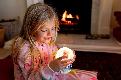 Front view of a happy young Caucasian girl sitting on the floor, holding a snow globe and smiling by the fireplace in her sitting room at Christmas time